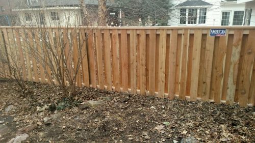 custom 6-foot tall wood board-on-board privacy fence with a top cap and 'American Fence' logo, seen from a bare ground area with shrubs and fallen leaves, with houses in the background.